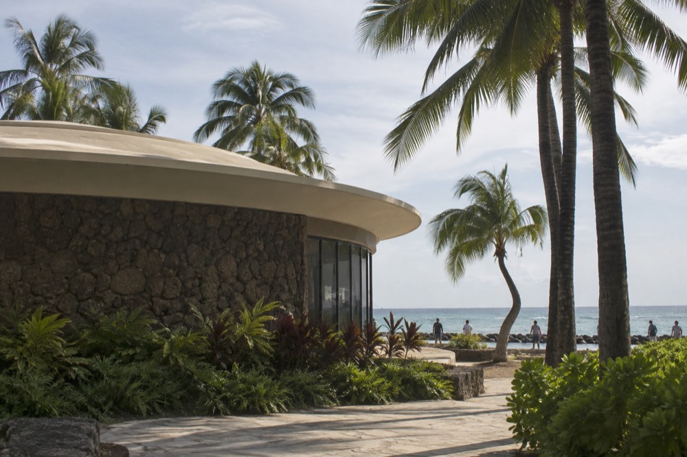 A low aquarium building beside palm trees near the Waikīkī shoreline, with the ocean visible behind it