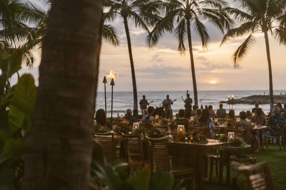 Long tables set for an outdoor Hawaiian feast under palm trees at dusk