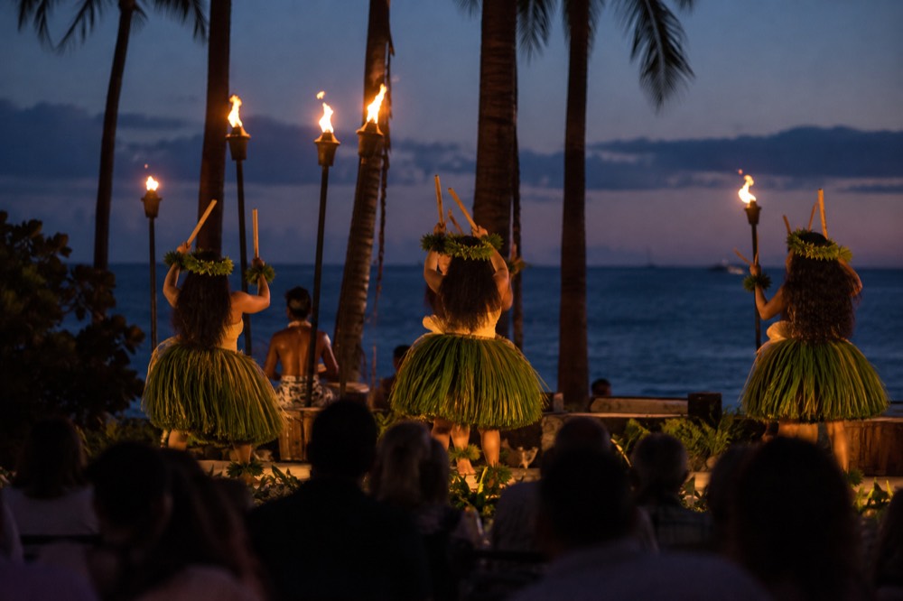 Performers in traditional attire dancing on an open-air stage lit by tiki torches at night