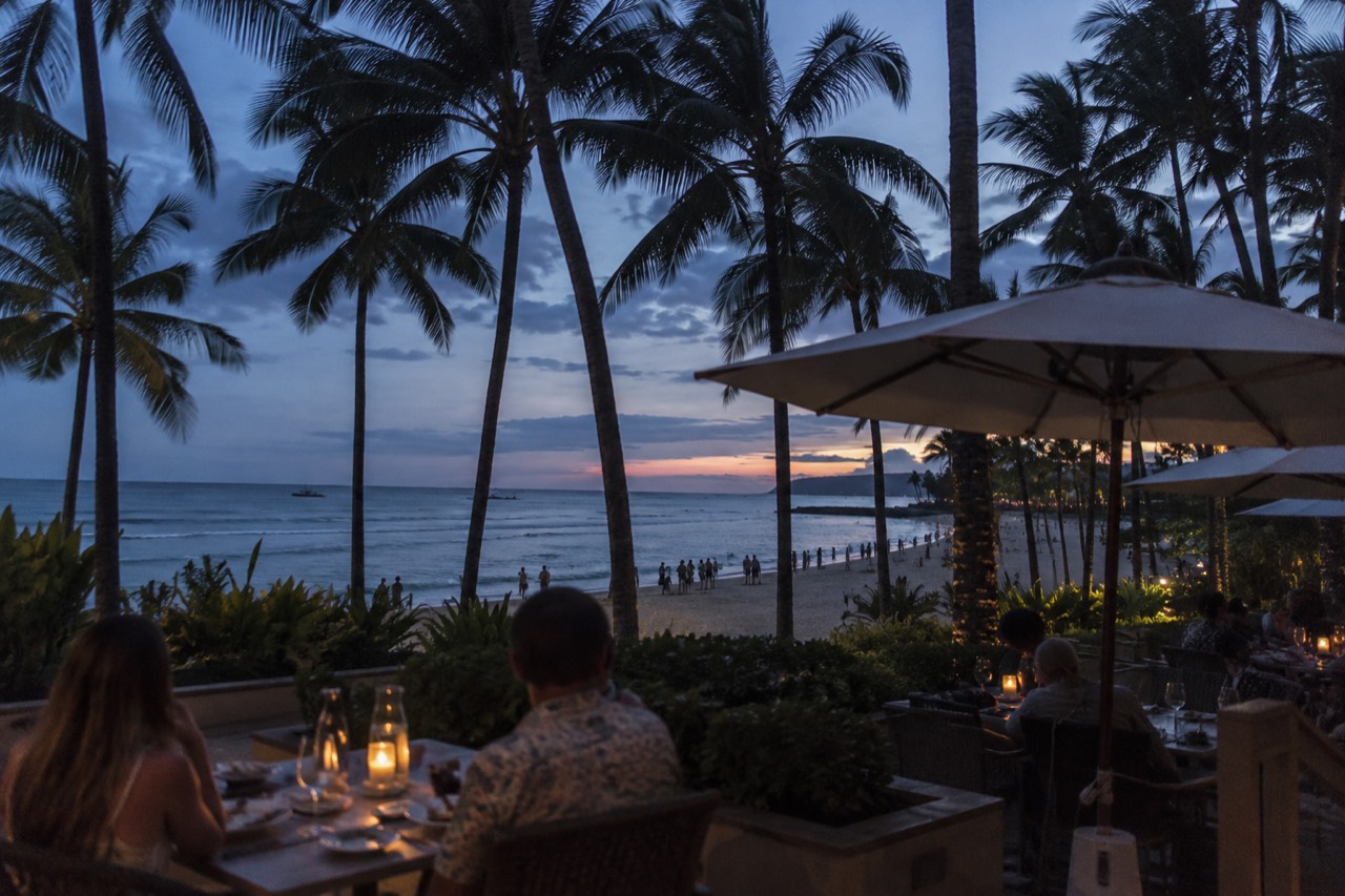 Outdoor seating at a Waikiki restaurant with palm trees and the beach visible beyond the tables
