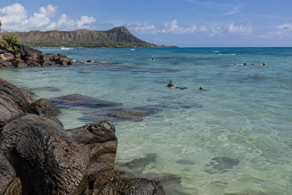 Snorkelers in calm shallow water above a sandy bottom near a rocky point, with green mountains in the background