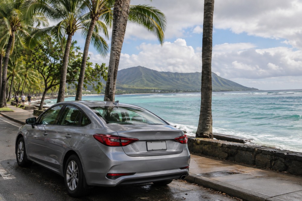 A rental car parked beside a palm-lined coastal road on Oahu with green mountains in the background