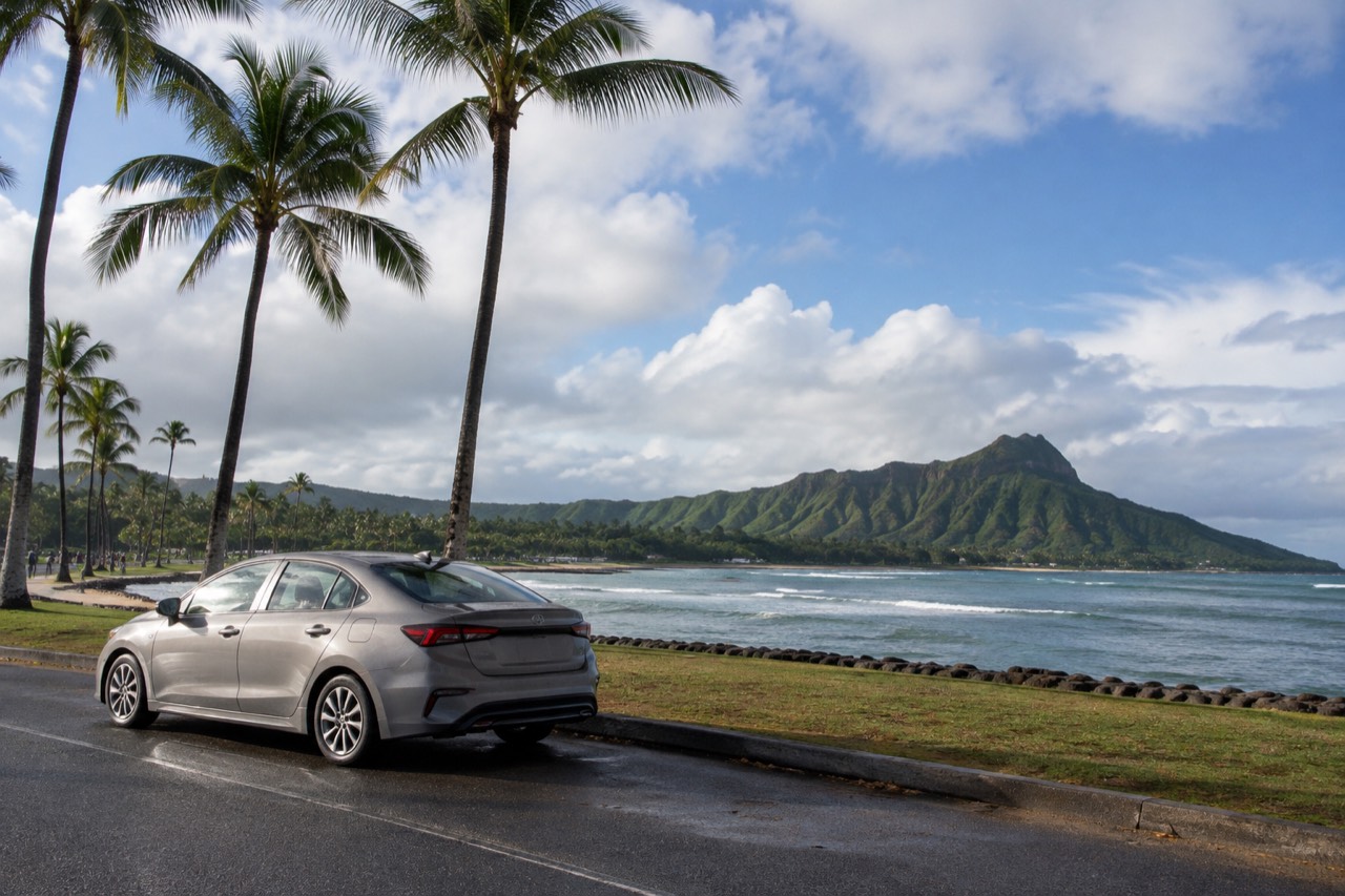 A rental car parked beside a palm-lined coastal road on Oahu with green mountains in the background