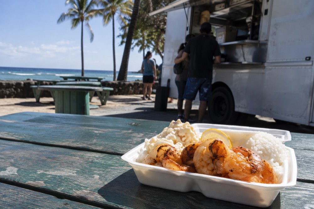 A picnic table beside a food truck with a plate lunch of shrimp and rice resting on it