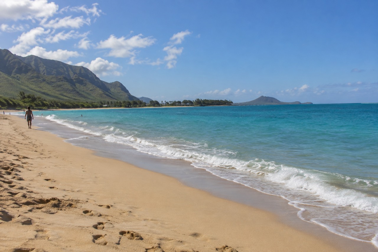 A wide sandy beach on Oahu with turquoise water and green mountains in the distance under a clear sky