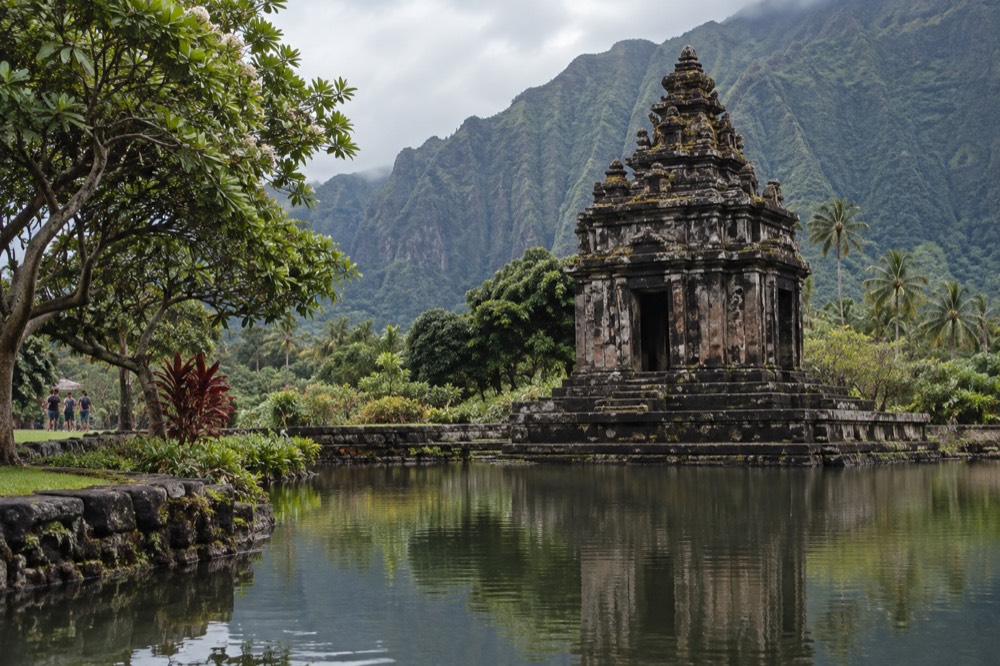 A stone temple reflected in a still pond with steep green mountain cliffs behind it