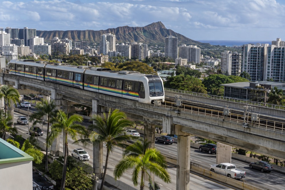 A Skyline train on an elevated guideway above a Honolulu highway with palm trees below
