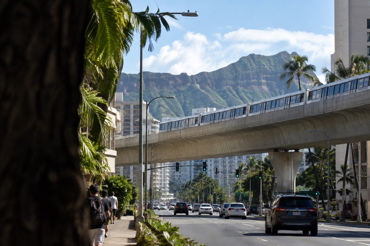 An elevated rail viaduct crossing above a Honolulu street with the Koʻolau mountains in the distance