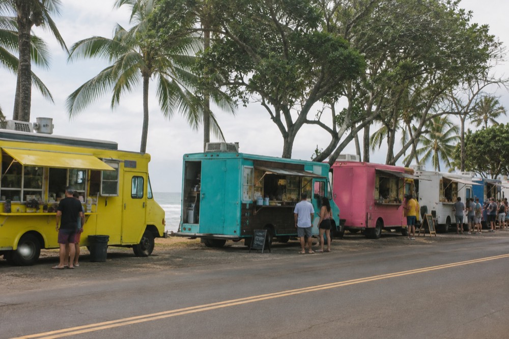 A row of colorful food trucks parked under trees near a beach town road