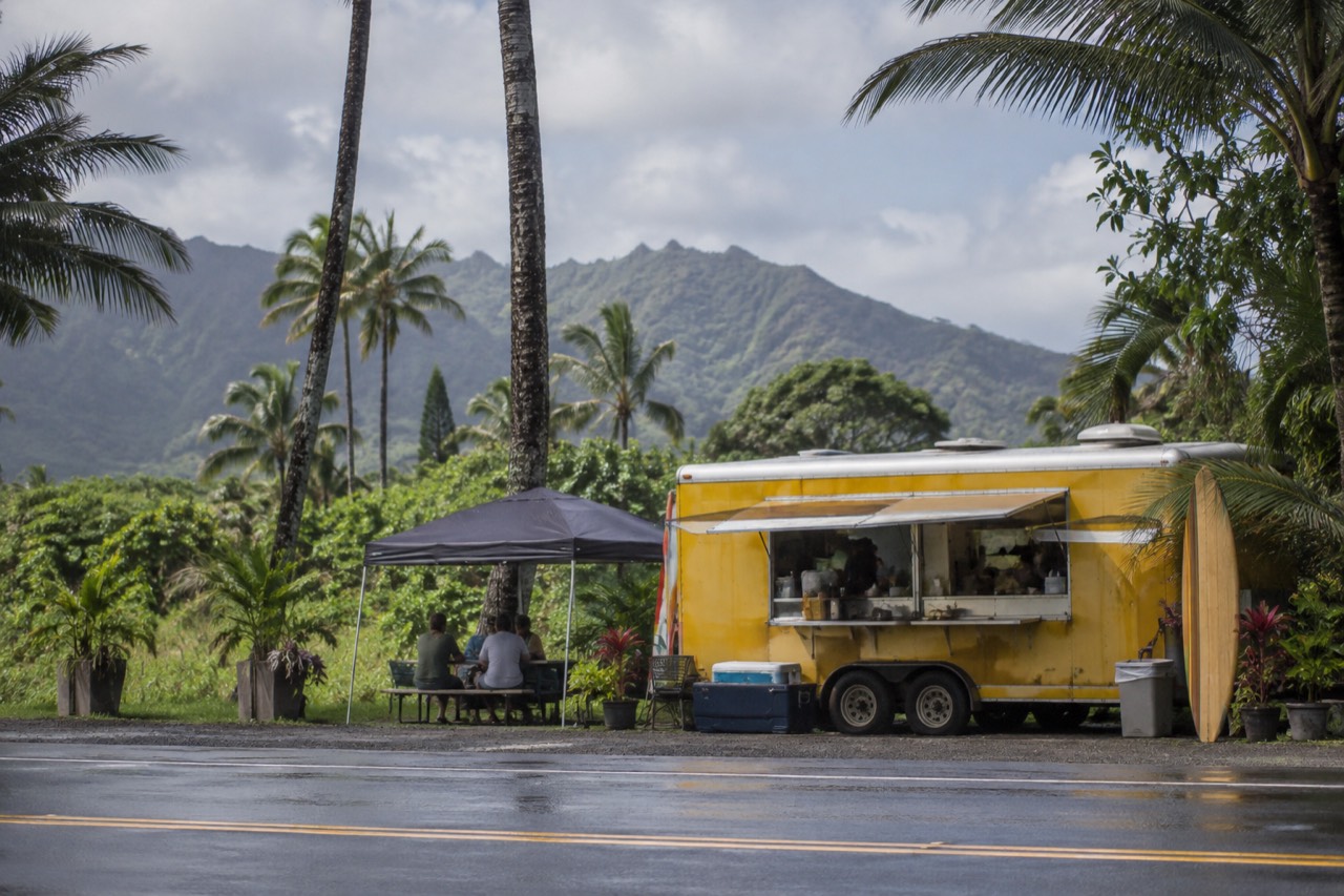 A food truck parked beside a two-lane highway with palm trees and green mountains behind it