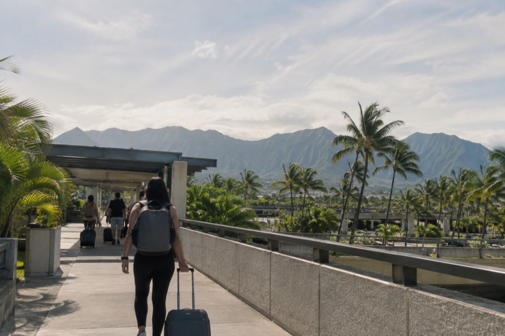 A visitor with a suitcase walking through an open-air airport walkway with palm trees and the Koʻolau mountains in the background