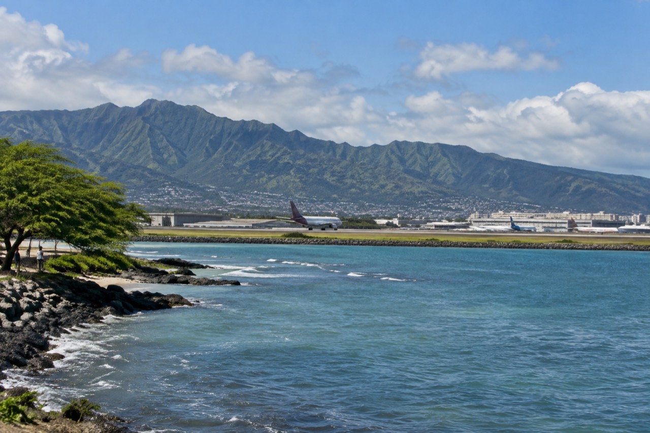 A wide view of the Honolulu airport approach with the Koʻolau mountain ridge rising behind the runways under scattered clouds