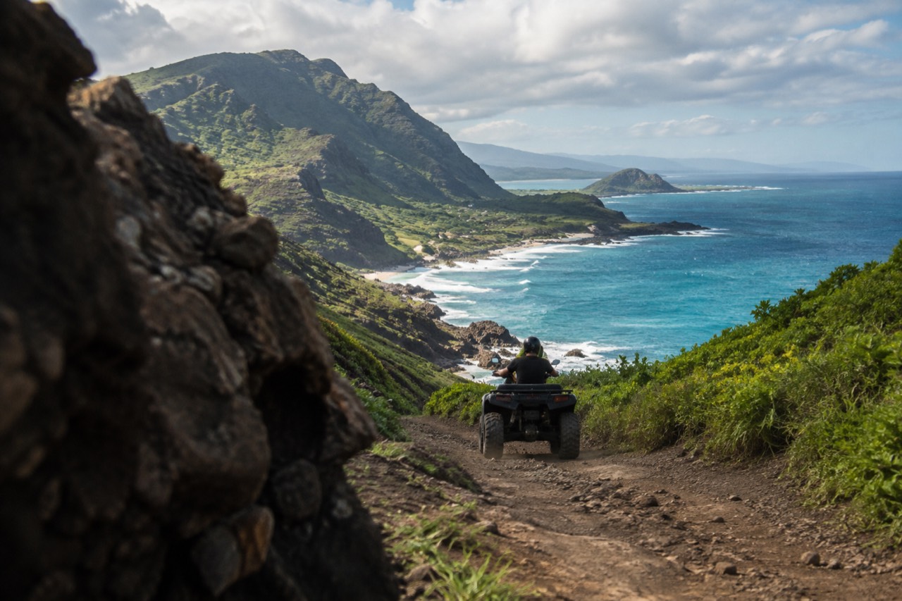 An all-terrain vehicle on a dirt track beside a green Oahu hillside near the ocean