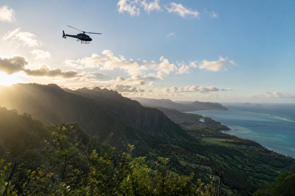 A helicopter flying above green Oahu ridgelines with the turquoise ocean in the distance