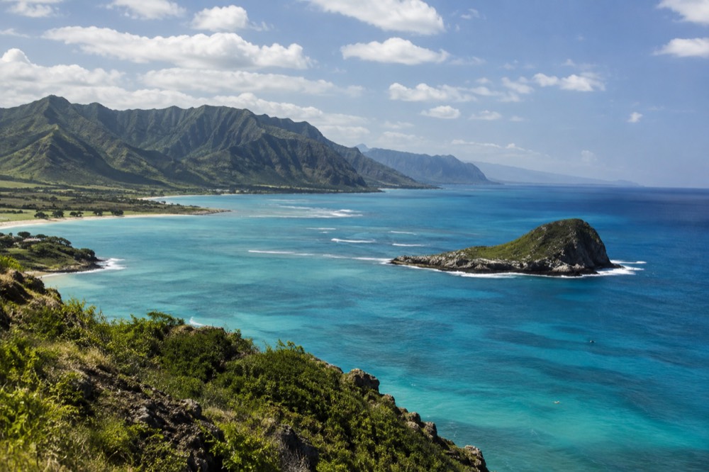 Aerial view of a turquoise bay with a small island offshore and steep green ridges behind it