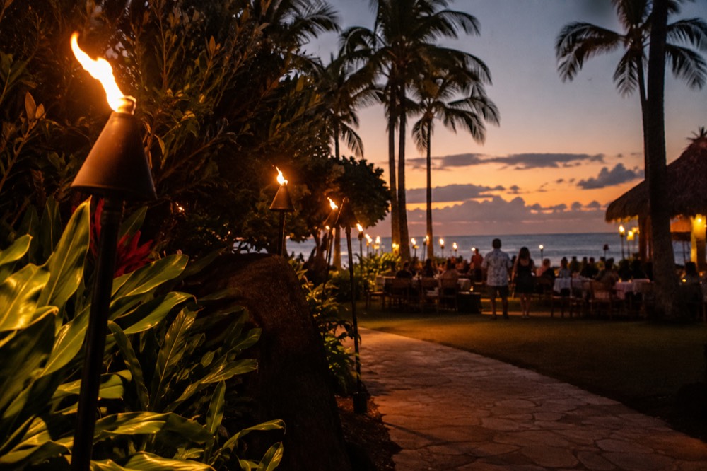 Dusk settling over an open-air luau ground on Oahu with tiki torches lit along a path