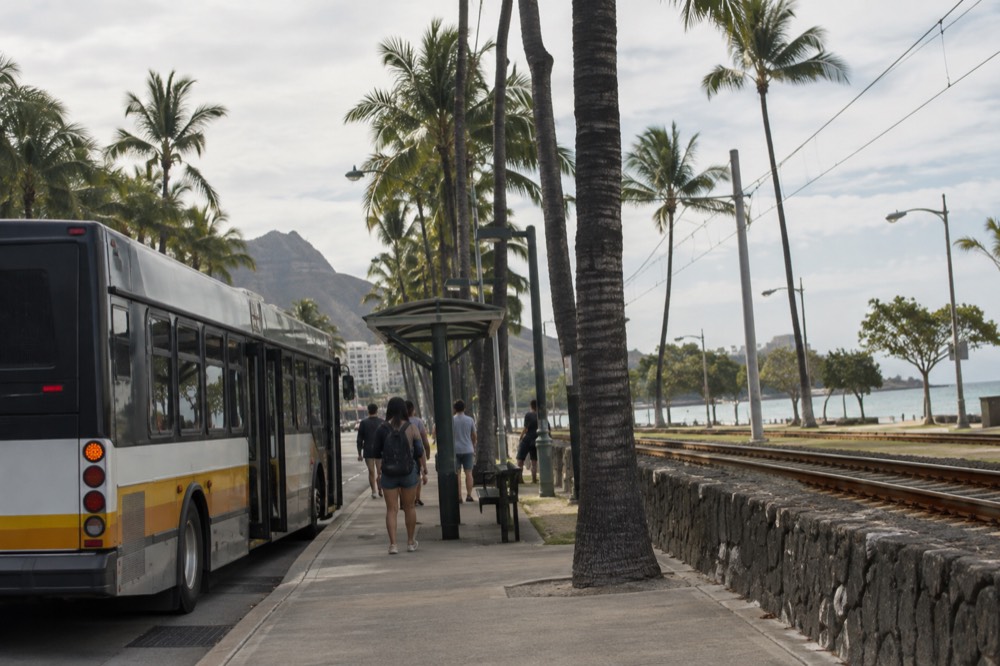 A Honolulu city bus at a stop with palm trees and a rail line nearby