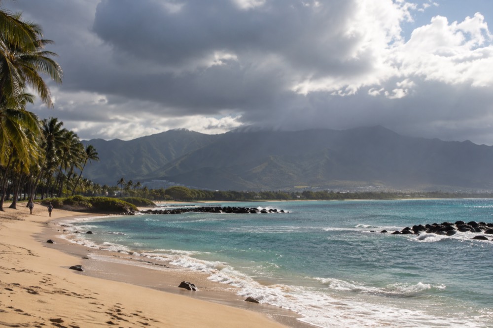 Dark rain clouds over the green ridges of the Koʻolau mountains, with sunlight breaking over the coastal plain below