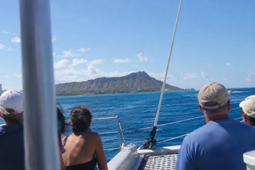 Visitors on a catamaran watching open water from the deck as the Oahu coastline slides past