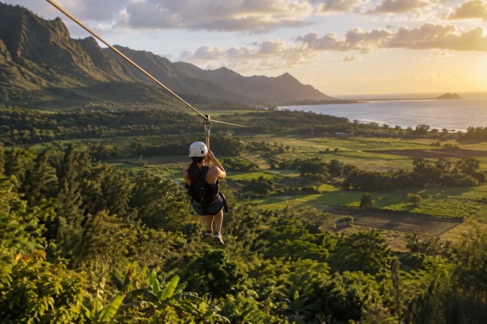 A person in a helmet and harness riding a zipline above tropical farmland on Oahu