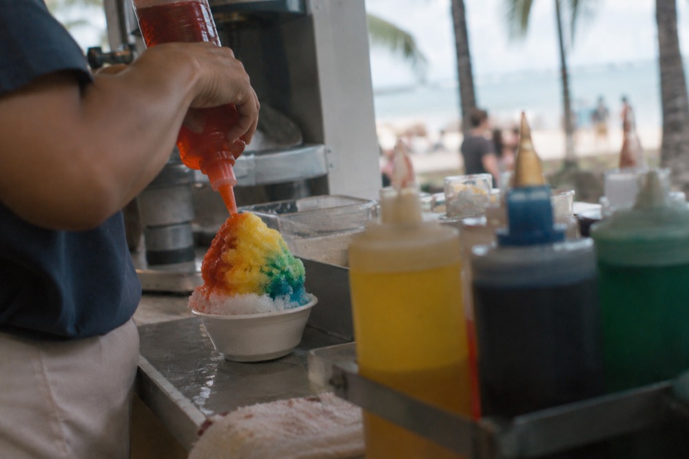 Close-up of finely shaved ice being drizzled with colorful syrup behind a shop counter