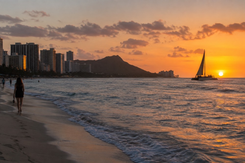A catamaran silhouetted against an orange sunset off Waikiki Beach with the skyline behind it