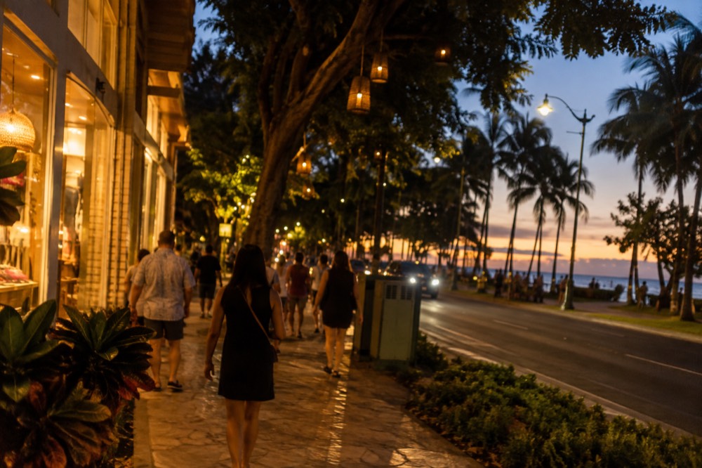 Kalakaua Avenue at night with lit shopfronts, banyan trees and pedestrians walking under string lights