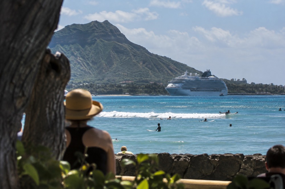 A cruise ship at anchor off the Oahu coastline with green mountains rising behind the water