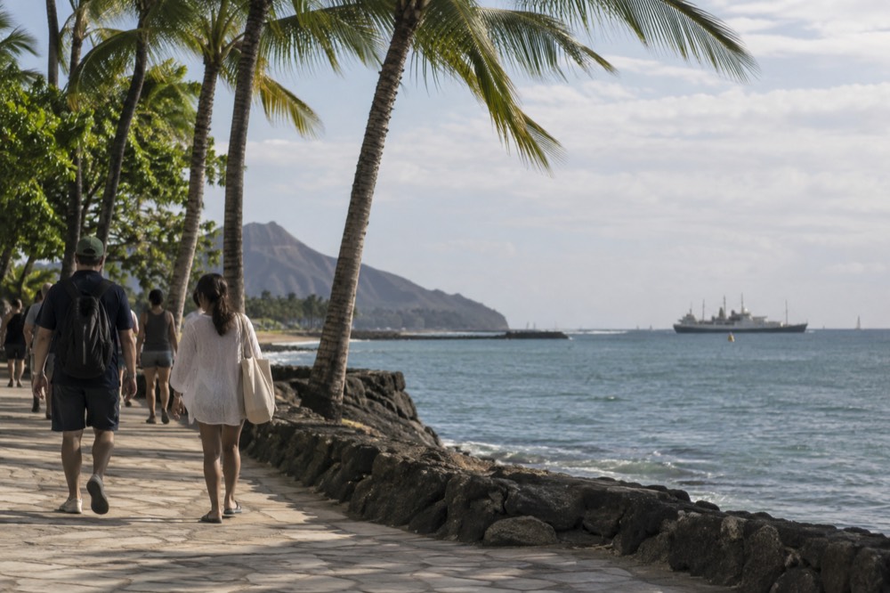 Passengers walking along a Honolulu waterfront with palm trees and an anchored ship in the background